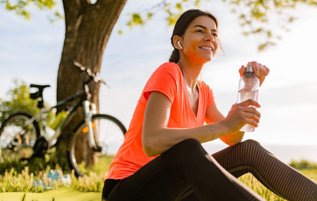 smiling beautiful woman drinking water in bottle doing sports in morning in park nature doing yoga on mat, pink fitness outfit, happy healthy lifestyle, music in earphones, bicycle on background
